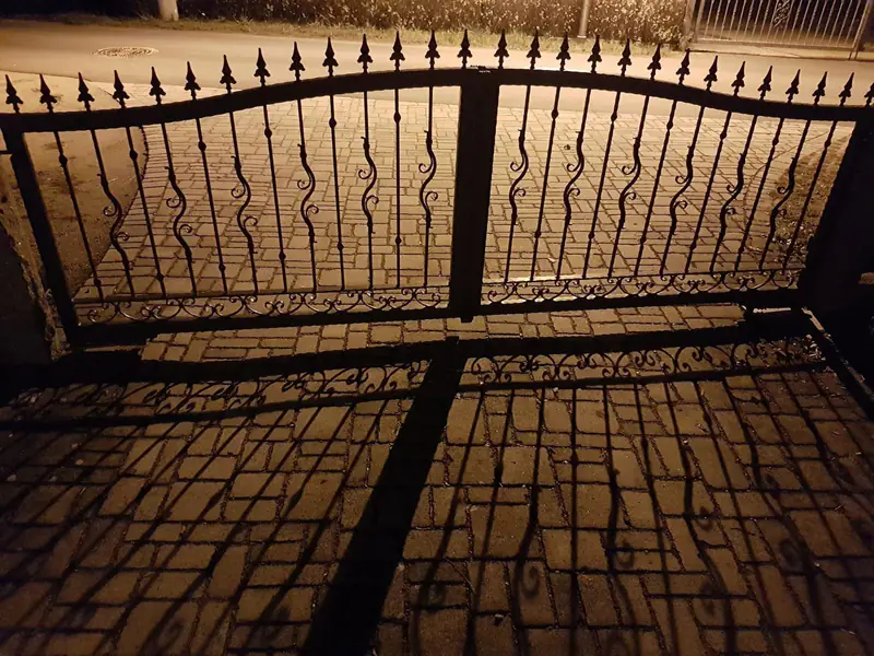 Zweiflügeliges schmiedeeisernes Bogentor mit Lilienspitzen, das nachts lange Schatten auf das Pflaster wirft