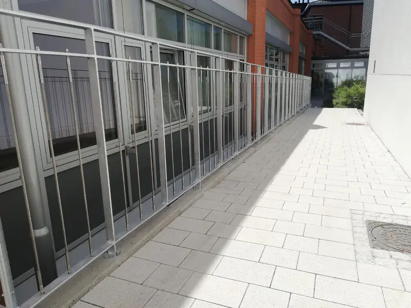 Feuerverzinktes Geländer mit senkrechter Seilfüllung entlang eines gepflasterten Gehwegs vor einem Klinkerbau