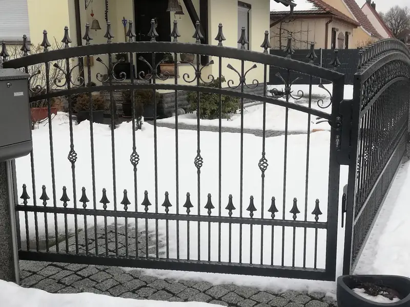 Schmiedeeiserne Pforte mit Bogen, Korbstäben und Lanzenspitzen vor einem verschneiten Vorgarten