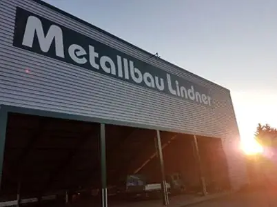 Werkhalle aus Trapezblech mit dem großen Schriftzug „Metallbau Lindner“, im Gegenlicht der Abendsonne